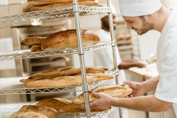 Handsome young baker putting fresh bread loaf on shelf at baking manufacture — Stock Photo