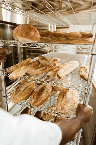 Tiro recortado do padeiro americano africano que conduz prateleiras do pão fresco na fabricação do cozimento — Fotografia de Stock