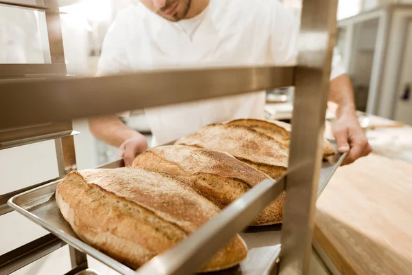 Cropped shot of baker putting trays of fresh bread on stand at baking manufacture — Stock Photo