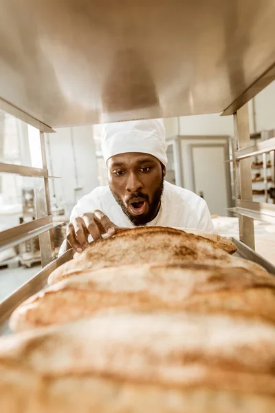 Shocked african american baker looking at fresh loaves of bread on baking manufacture — Stock Photo