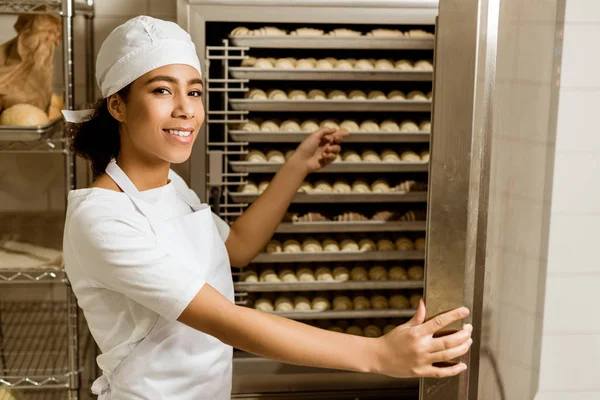 Улыбающаяся женщина-пекарь, указывающая на тесто внутри промышленной печи при выпечке — стоковое фото