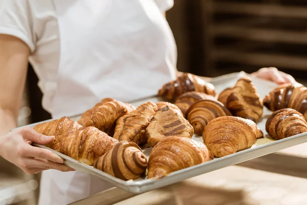 Cropped shot of female baker holding tray with fresh croissants on baking manufacture — Stock Photo