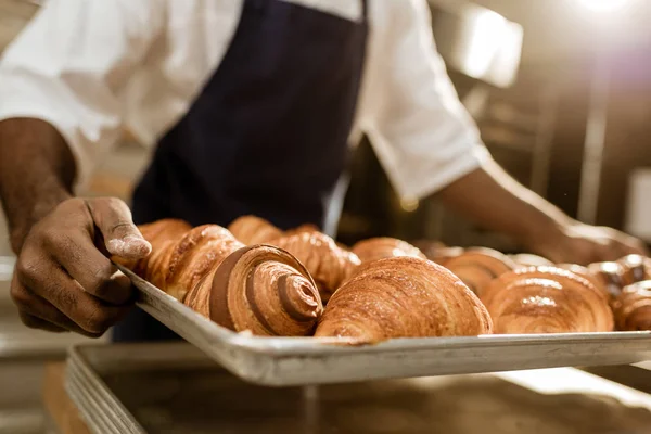 Cropped shot of baker holding tray with croissants on baking manufacture — Stock Photo