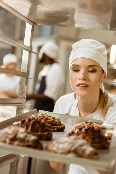 Привлекательная женщина-пекарь смотрит на поднос свежеиспеченных круассанов на выпечке — стоковое фото
