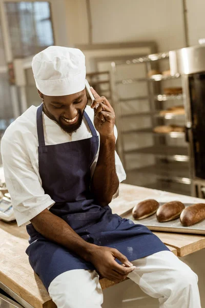 Panadero afroamericano joven con las manos cubiertas de harina hablando por teléfono mientras está sentado en la mesa de trabajo - foto de stock