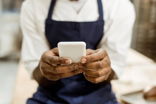 Recortado tiro de panadero afroamericano con las manos cubiertas de harina usando teléfono inteligente - foto de stock