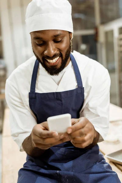 Panadero afroamericano joven con las manos cubiertas de harina usando teléfono inteligente - foto de stock