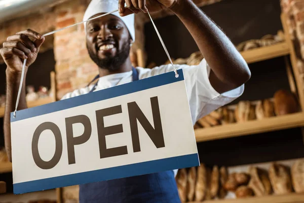 Happy african american baker holding open sign at pastry store — Stock Photo