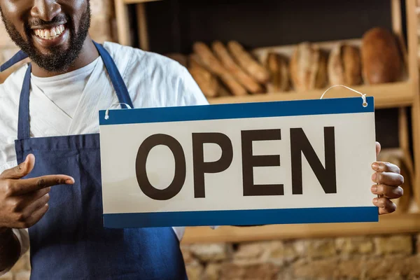 Colpo ritagliato di panettiere afroamericano che punta il cartello aperto al negozio di pasticceria — Foto stock