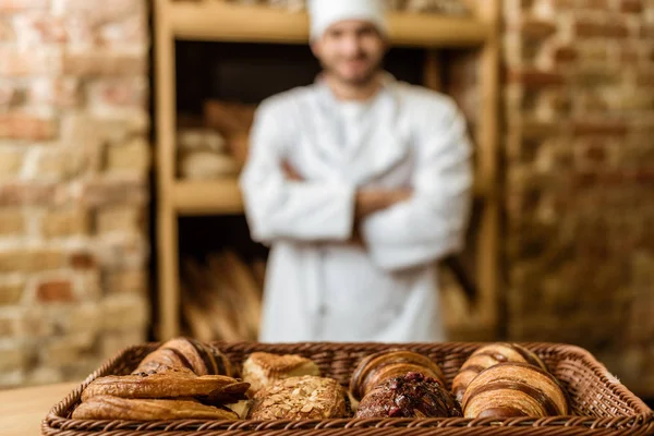 Blurred baker with crossed arms standing at pastry store with basket of croissants on foreground — Stock Photo