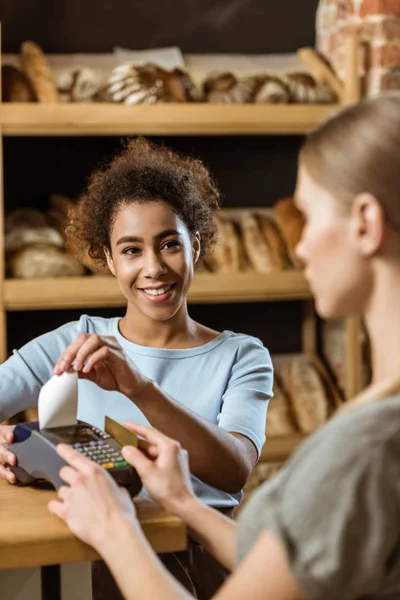 Young cashier with pos terminal receiving purchase from client at pastry store — Stock Photo