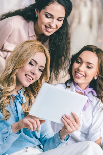 Young smiling bride and bridesmaids choosing dresses and looking at tablet in wedding fashion shop — Stock Photo