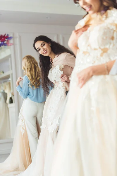 Joven novia sonriente y damas de honor elegir vestidos en la tienda de moda de boda - foto de stock
