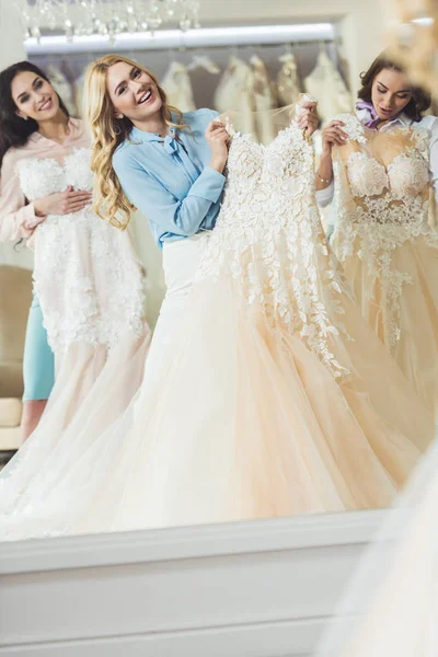 Happy bride and bridesmaids trying on dresses by mirror in wedding fashion shop — Stock Photo