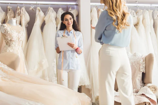 Красивая невеста и женщина портной обсуждают дизайн одежды в свадебном салоне — стоковое фото
