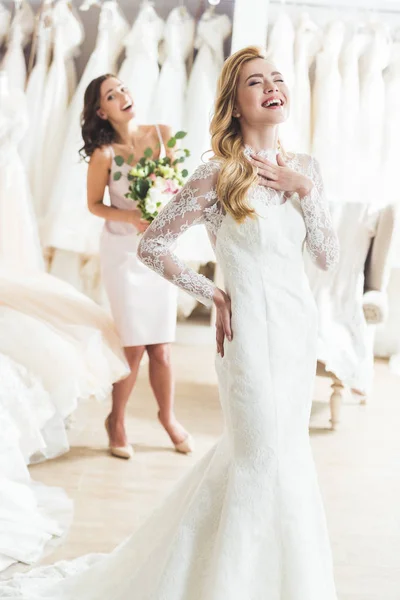 Novia sonriente y dama de honor en la tienda de moda de bodas - foto de stock