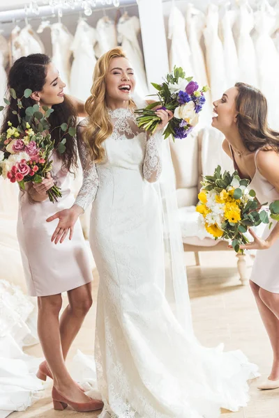 Joven novia sonriente y damas de honor con ramos en la tienda de moda de boda - foto de stock