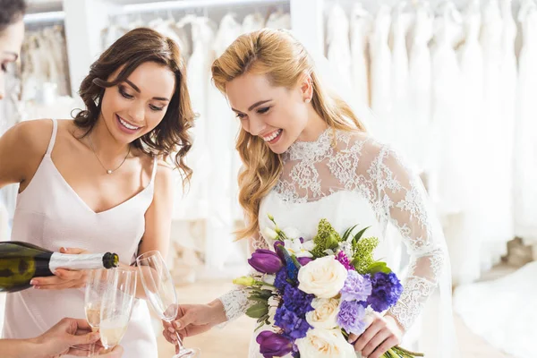 Novias jóvenes sosteniendo copas de champán y botella en la tienda de moda de boda - foto de stock