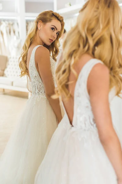 Jolie femme portant une robe de mariée par miroir dans un atelier de mariage — Photo de stock