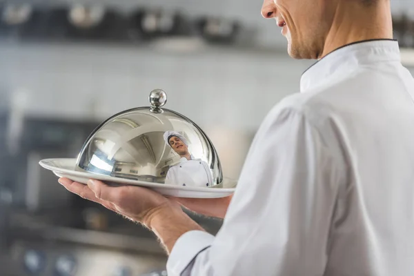 Imagem cortada de belo chef segurando prato com tampa na cozinha do restaurante — Fotografia de Stock