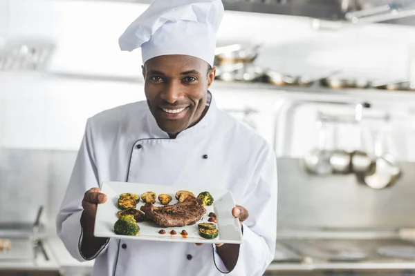 Chef afroamericano feliz celebración plato con filete cocido y verduras en la cocina del restaurante - foto de stock