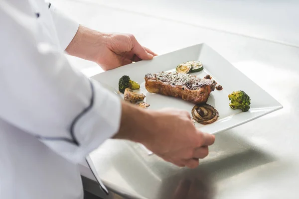 Обрезанный образ шеф-повара, держащего тарелку со стейком и овощами на кухне ресторана — стоковое фото