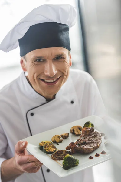 Bonito chef segurando prato com bife cozido na cozinha do restaurante — Fotografia de Stock