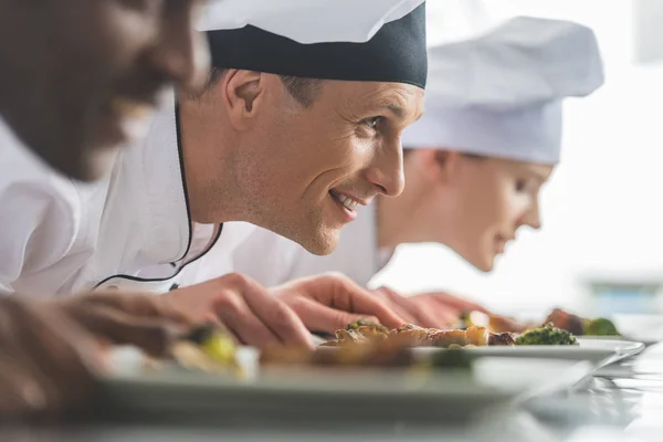 Chefs multiétnicos sonrientes olfateando comida cocinada en la cocina del restaurante — Stock Photo