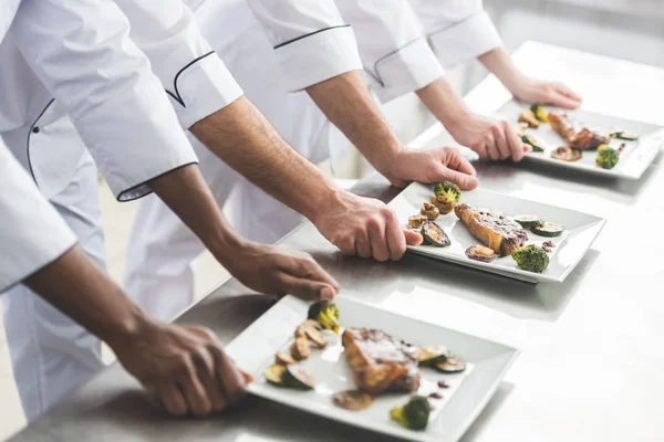 Cropped image of multicultural chefs with plates with steaks at restaurant kitchen — Stock Photo