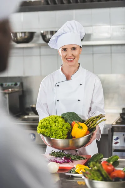 Улыбающийся шеф-повар держит миску с овощами и смотрит в камеру на кухне ресторана — стоковое фото