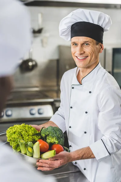 Chef souriant tenant bol avec des légumes et en regardant collègue à la cuisine du restaurant — Photo de stock