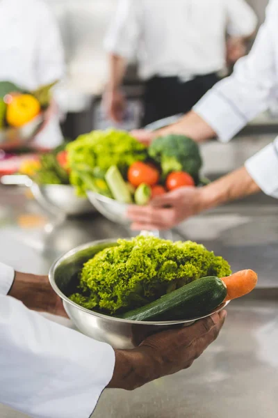 Imagen recortada de chefs multiétnicos sosteniendo cuencos con verduras en la cocina del restaurante - foto de stock