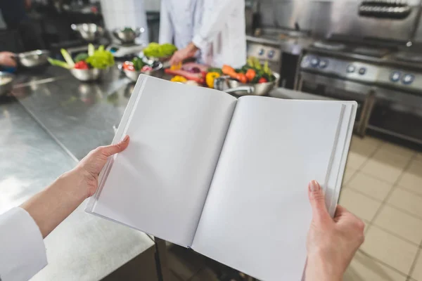 Abgeschnittenes Bild von Koch mit offenem Rezeptbuch in Restaurantküche — Stockfoto