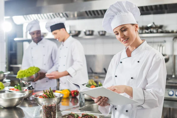 Attractive chef reading recipe in recipe book at restaurant kitchen — Stock Photo