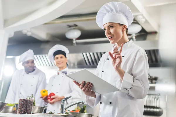 Chef atraente segurando livro de receitas e mostrando gesto ideia na cozinha do restaurante — Fotografia de Stock