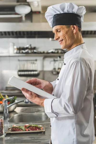 Vista laterale del bel chef lettura libro di ricette al ristorante cucina — Foto stock