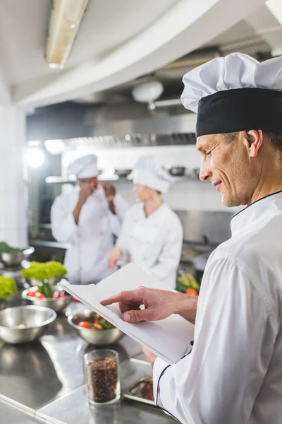 Vista lateral del chef apuntando al libro de recetas en la cocina del restaurante - foto de stock