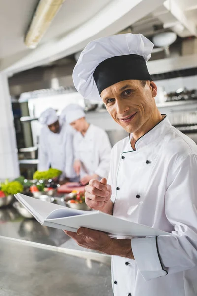 Chef sorrindo segurando livro de receitas e olhando para a câmera na cozinha do restaurante — Fotografia de Stock