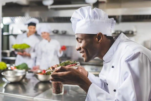 Sonriente chef afroamericano olfateando carne cruda con hierbas en la cocina del restaurante - foto de stock