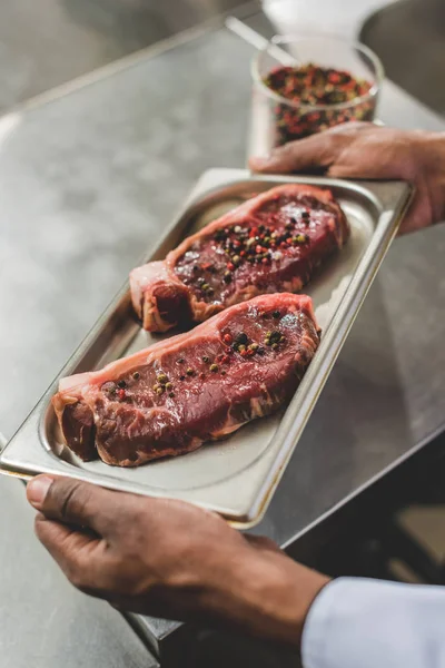 Abgeschnittenes Bild eines afrikanisch-amerikanischen Kochs mit Tablett mit rohem Fleisch in der Restaurantküche — Stockfoto