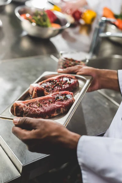 Abgeschnittenes Bild eines afrikanisch-amerikanischen Kochs mit Tablett mit rohem Fleisch in der Restaurantküche — Stockfoto