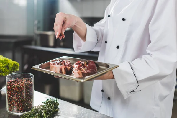 Cropped image of chef adding spices to raw steaks at restaurant kitchen — Stock Photo