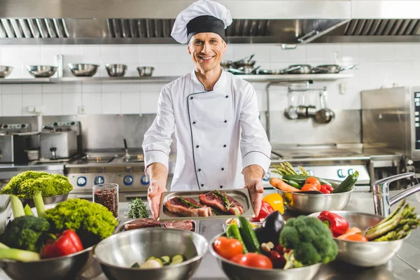 Lächelnder Koch nimmt Tablett mit rohem Fleisch in Restaurantküche — Stockfoto