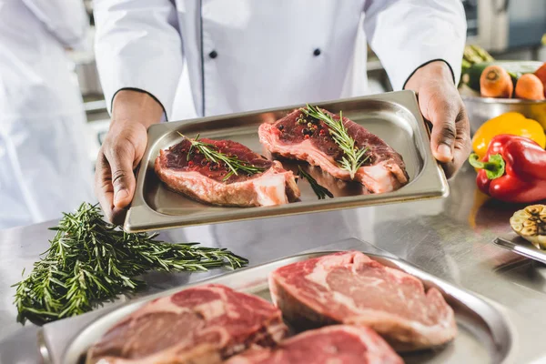 Image recadrée du chef afro-américain tenant un plateau avec des steaks crus à la cuisine du restaurant — Photo de stock