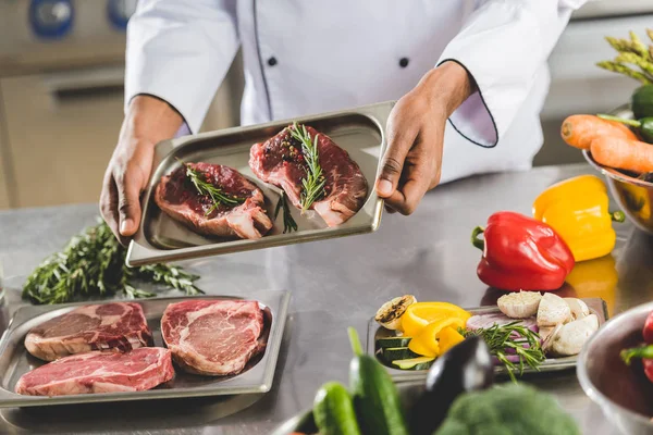 Abgeschnittenes Bild eines afrikanisch-amerikanischen Kochs mit Tablett mit rohem Fleisch in der Restaurantküche — Stockfoto