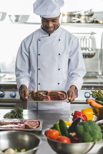 Улыбающийся красивый африканский шеф-повар держит поднос с сырым мясом на кухне ресторана — стоковое фото