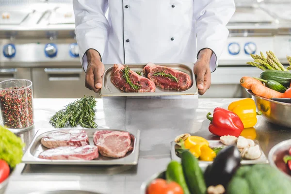 Image recadrée du chef afro-américain tenant un plateau avec de la viande non cuite au restaurant cuisine — Photo de stock