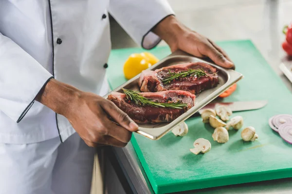 Imagen recortada del chef afroamericano sosteniendo bandeja con carne cruda en la cocina del restaurante - foto de stock