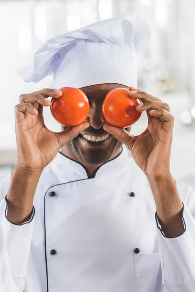 Guapo afroamericano chef cubriendo los ojos con tomates en restaurante cocina - foto de stock