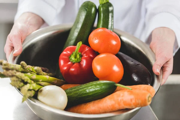Imagen recortada de chef sosteniendo tazón de verduras orgánicas maduras en la cocina del restaurante - foto de stock